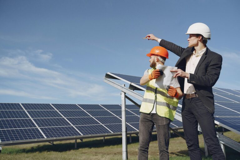 Young businessman in formal outfit and foreman in yellow vest looking away while discussing working process against solar panels